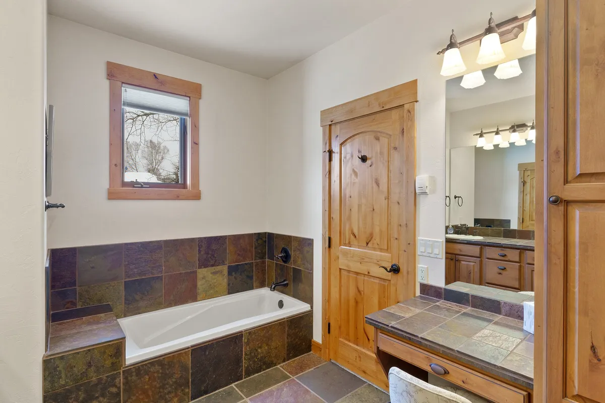 A tranquil bathroom scene with a freestanding bathtub, slate tile, wooden cabinetry, and a large mirror. Natural light filters through the window, creating a cozy and spa-like environment ideal for unwinding after outdoor activities.