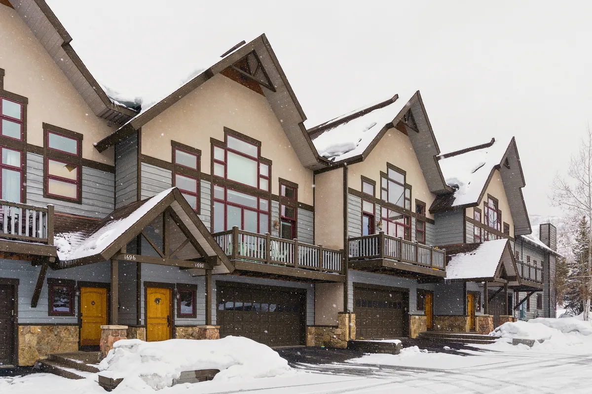 The image shows snow-covered rooftops and decorative balconies of Alpine Vista Chalet, with wooden doors and a winter landscape. The warm glow from the windows contrasts beautifully with the snowy exterior, evoking the anticipation of winter sports and cozy evenings after a day on the slopes.