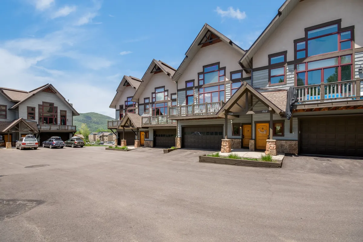 A broad view of the townhouse exteriors with multiple balconies, a driveway, and parked cars. The communal outdoor spaces are bathed in sunset light, suggesting lively family gatherings, barbecues, and outdoor games.